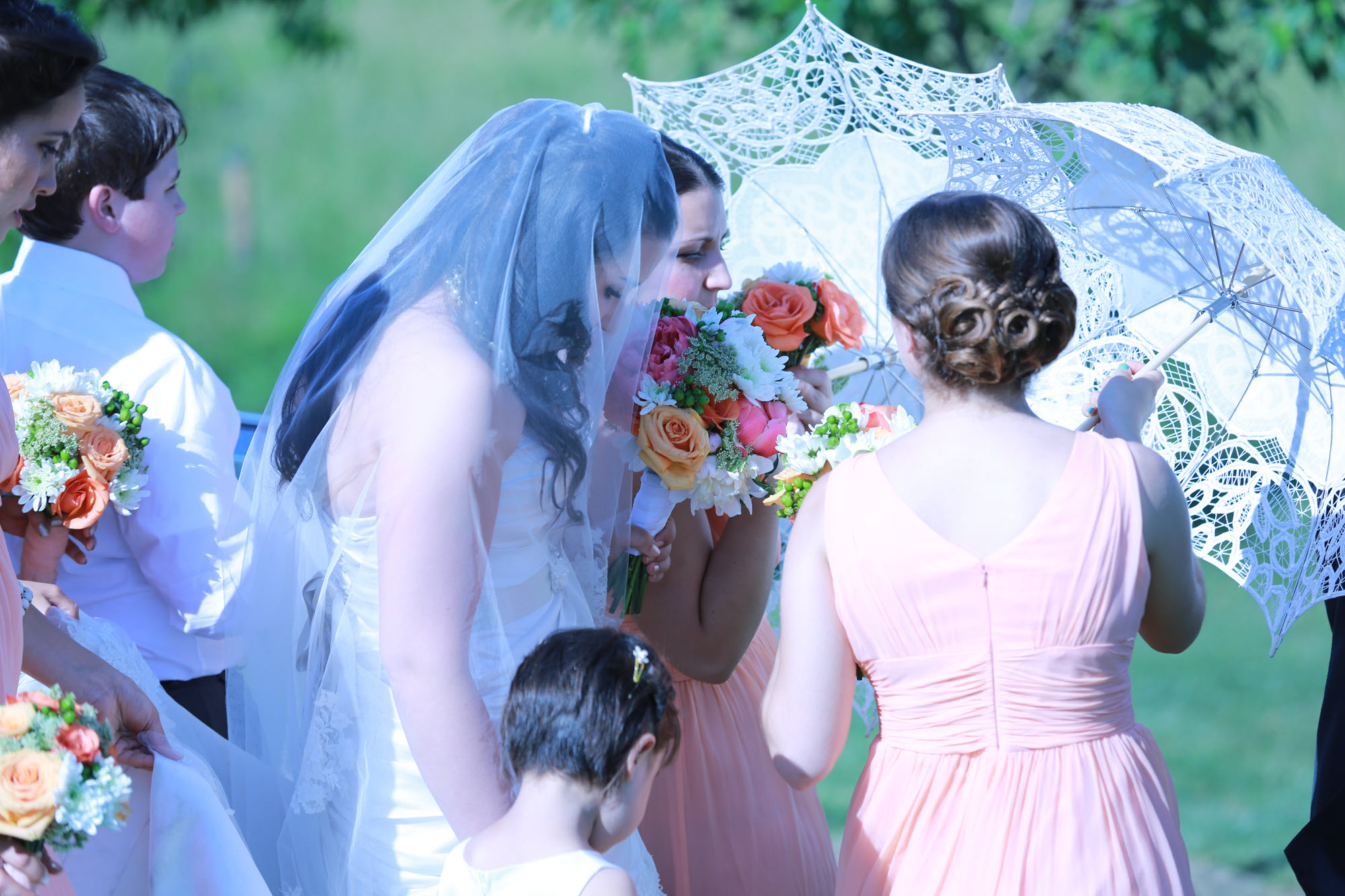 Bridesmaids holding colorful bouquets, shaded by a stylish umbrella under the summer sun at a wedding celebration.