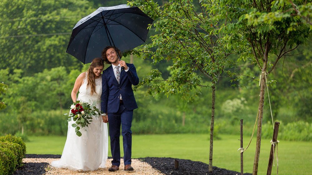 Bridal couple holding hands under an umbrella in the rain, with the bride clutching a bouquet.