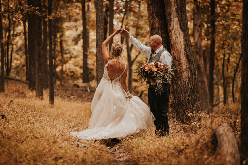 A joyful bride twirls in her wedding dress while the groom playfully holds the bouquet, both sharing a blissful moment in a serene park.