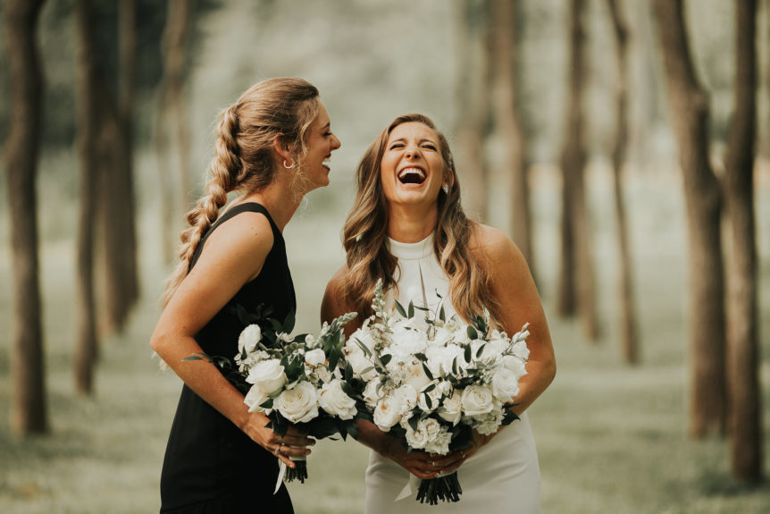 Bride and Bridesmaid Laughing in Wooded Area During All-Weekend Wedding Celebration with Bouquets