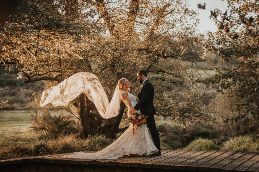 Joyful Bridal Couple Standing on Bridge with Veil Blowing in the Wind