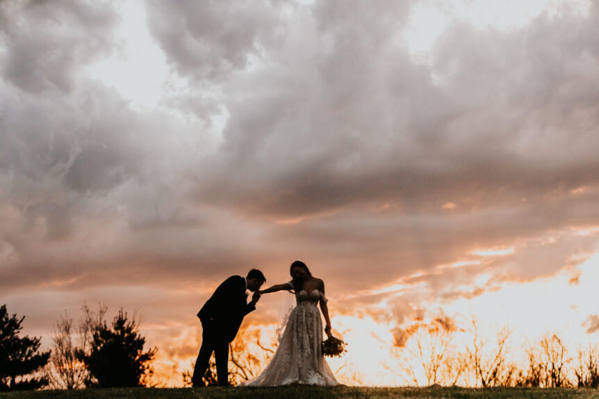 groom kissing bride's hand against sunset background in a field