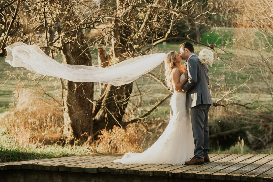 Newlyweds sharing a kiss on the wooden bridge at Zion Springs all-inclusive wedding venue, with a flowing veil and scenic countryside in the background.